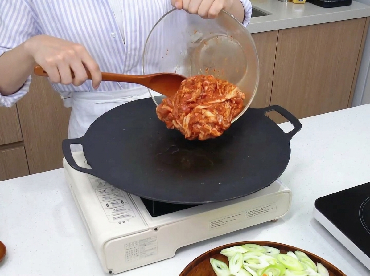 Marinierter roter Schweinebauch wird mit einem Holzspatel aus einer Glasschüssel in eine heiße, schwarze Pfanne gegeben.