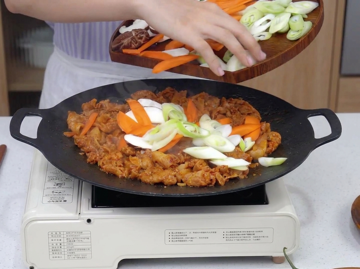 Verschiedenes geschnittenes Gemüse wie Zwiebeln und Karotten wird zum brutzelnden Schweinefleisch in die Pfanne gegeben.