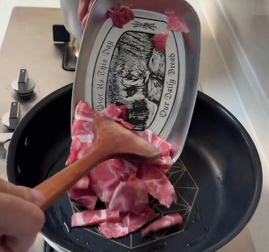 Dünne Scheiben rohes Schweinefleisch werden mit einem Holzspatel von einem Metalltablett in eine heiße Pfanne gegeben.