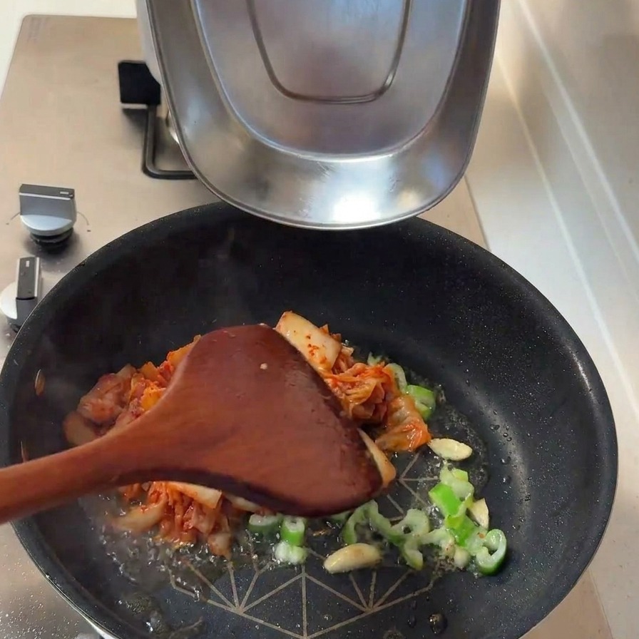 Fermentiertes Kimchi und dicke weiße Zwiebelscheiben werden zu den angeschwitzten Aromaten in die Pfanne gegeben.