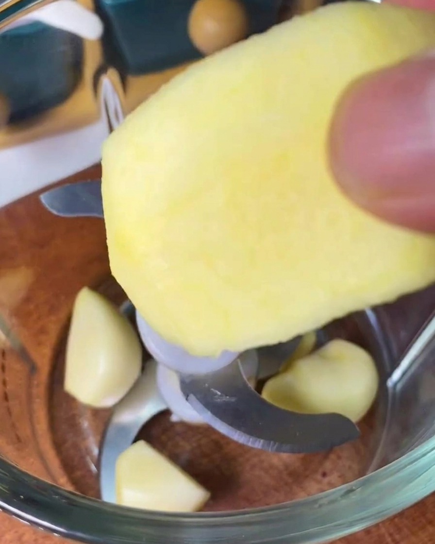 Ein Apfelstück wird in einen Mixer gegeben, in dem sich bereits geschälte Knoblauchzehen befinden.