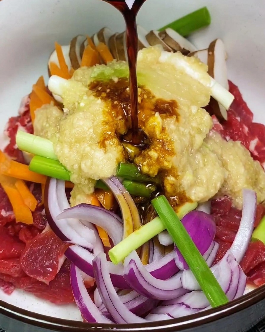 Dunkle Marinade wird über eine Mischung aus rohem Rindfleisch, Gemüse und hellem Fruchtpüree in einer weißen Schüssel gegossen.