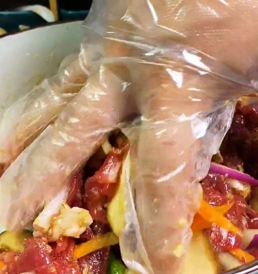 Eine Hand mit Handschuh mischt rohes Rindfleisch, Zwiebeln, Karotten und Pilze in einer dunklen Marinade gründlich durch.