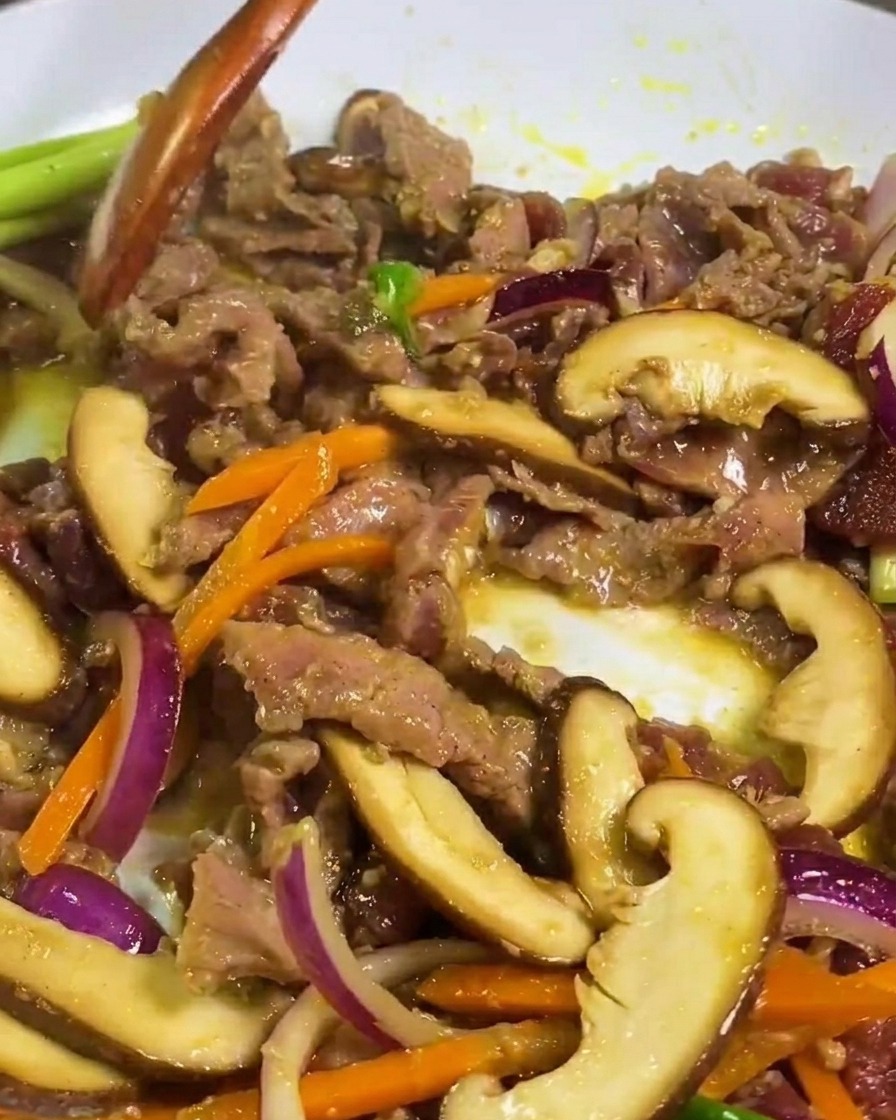 Dünne Fleischscheiben, Shiitake-Pilze und Karotten werden mit einem Holzspatel in einer Pfanne gebraten.