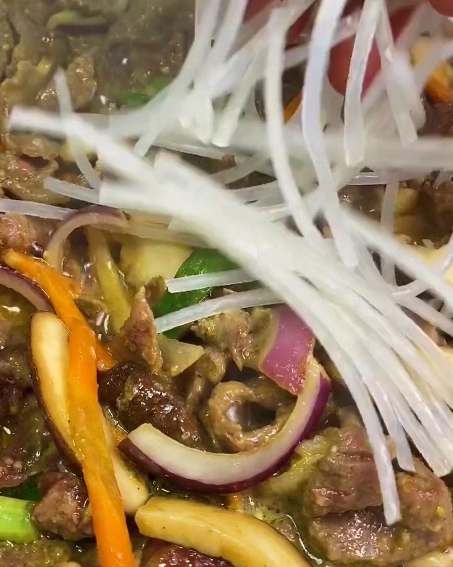 Eine Hand gibt eingeweichte, durchsichtige Süßkartoffel-Glasnudeln in die Pfanne zu Fleisch und Gemüse.
