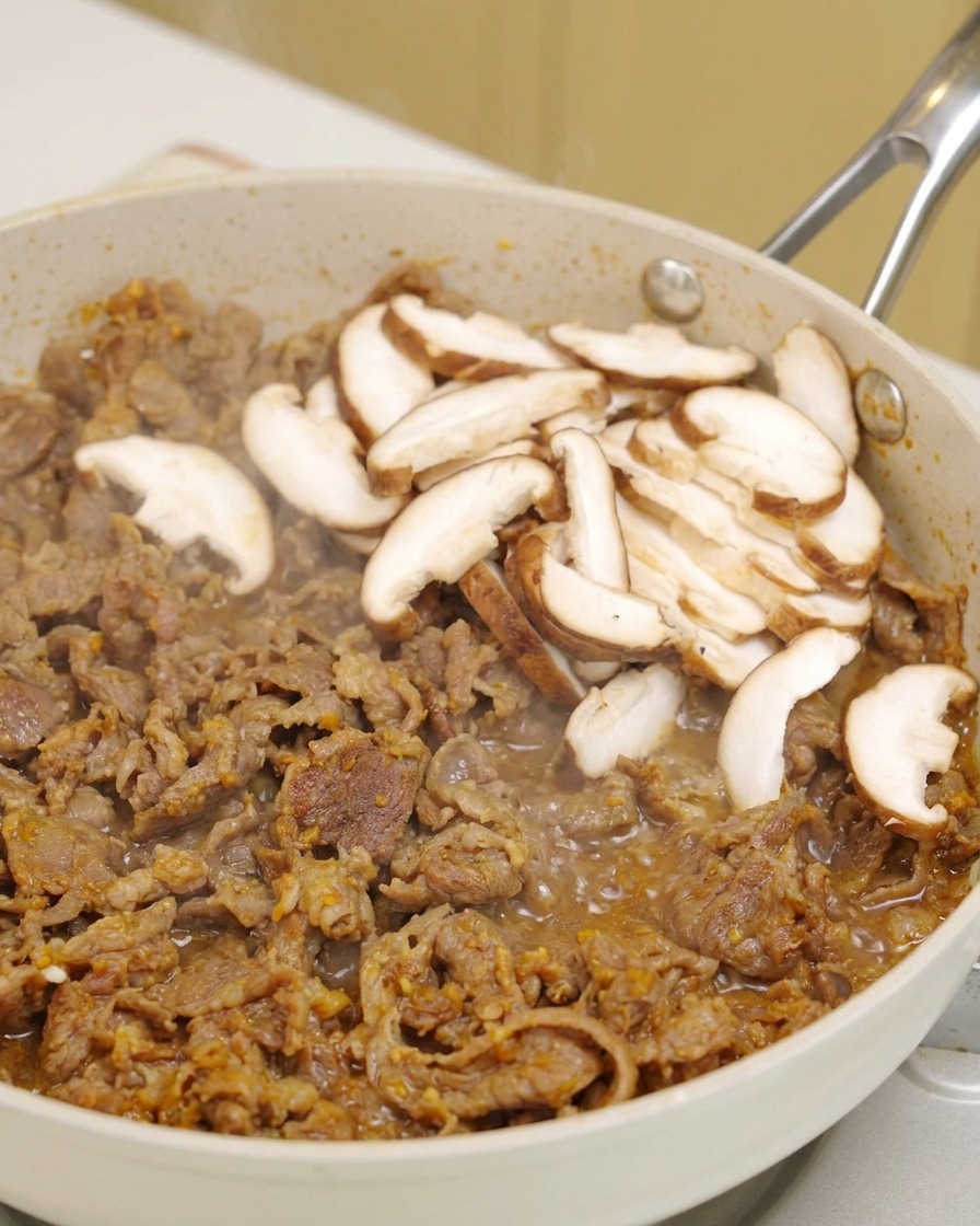 Frische Shiitake-Pilze werden auf das brutzelnde Fleisch in einer Pfanne gelegt.