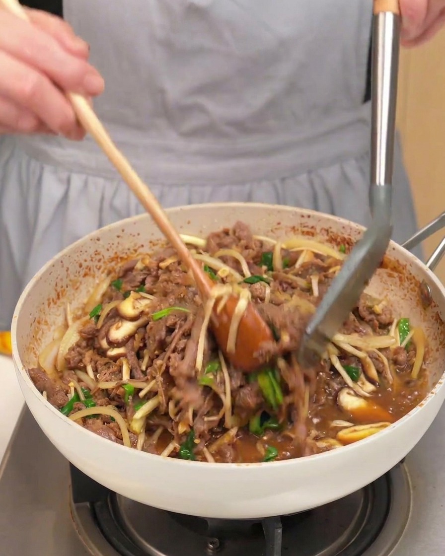 Eine Pfanne voll mit gegartem Fleisch, Pilzen, Zwiebeln und Frühlingszwiebeln wird mit einem Holzlöffel gewendet.