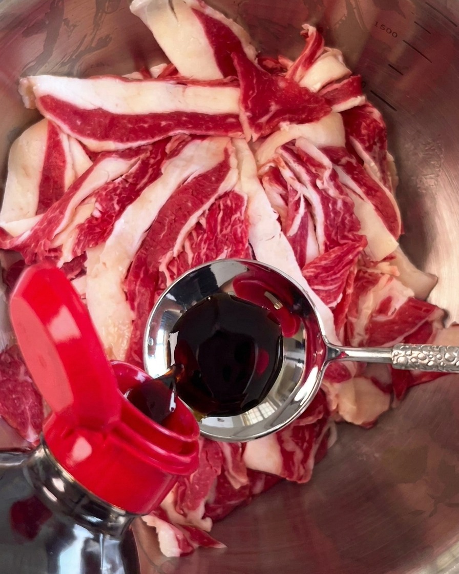 Dunkle Sojasauce wird mit einem Metalllöffel über das rohe Rindfleisch gegossen.