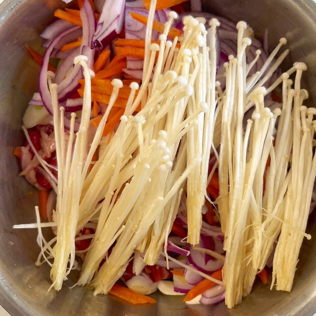 Weiße Enoki-Pilze und gehackter Knoblauch liegen auf dem Fleisch und dem Gemüse.