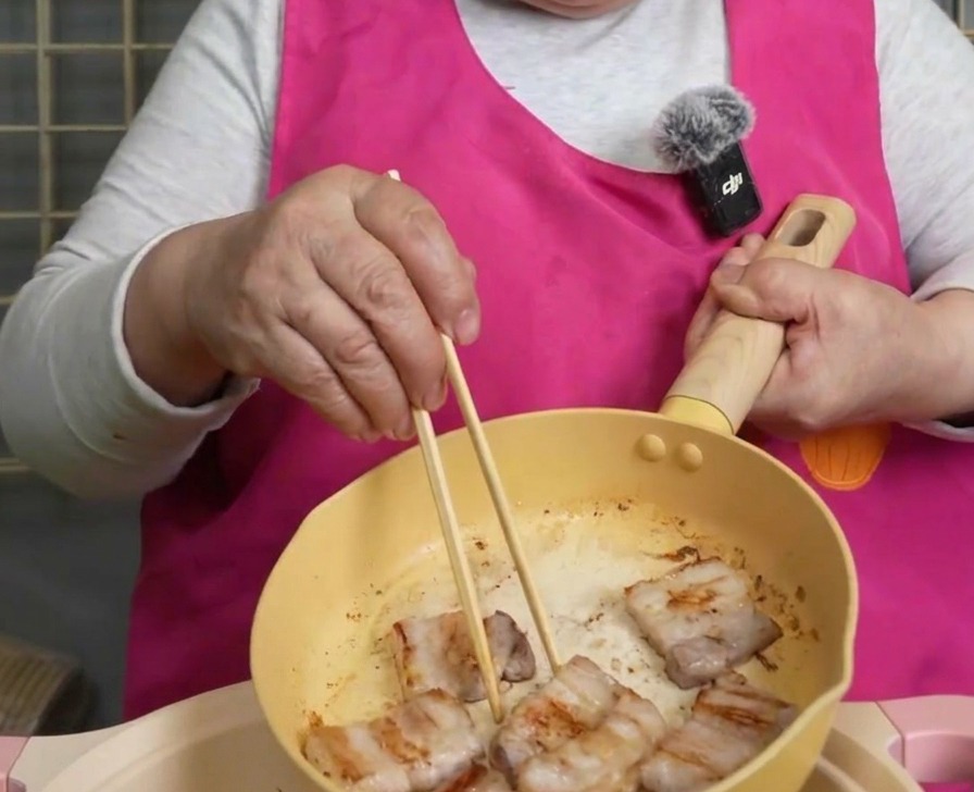 Gebraunte und knusprige Schweinebauchscheiben werden mit Holzstäbchen aus einer gelben Pfanne gehoben.