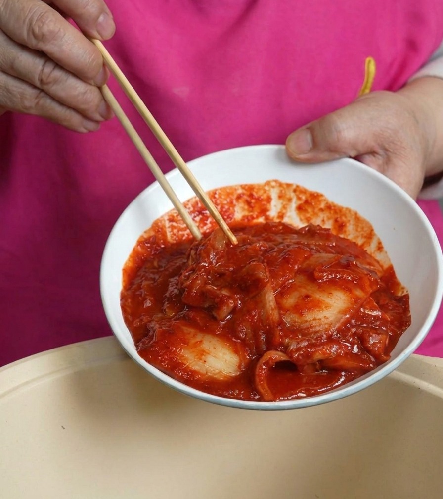 Eine weiße Schale mit leuchtend rotem, würzigem Kimchi wird über den Kochtopf gekippt.