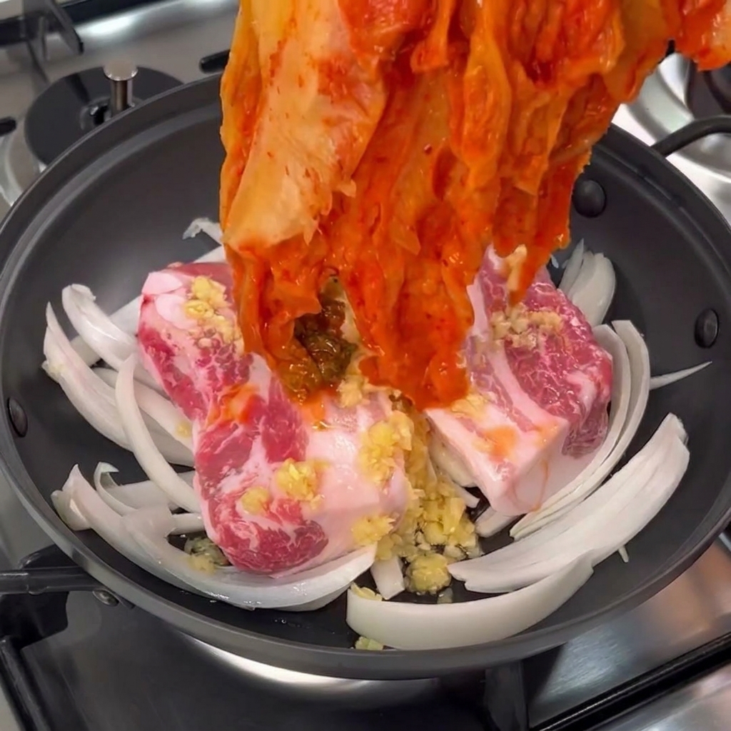 Eine Hand legt ein großes Bündel roten, gereiften Kimchi auf den Schweinebauch im Topf.