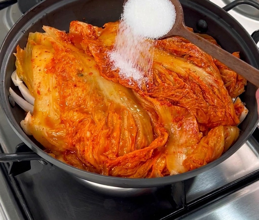 Weißer Zucker wird von einem Holzlöffel auf den roten Kimchi im Kochtopf gestreut.