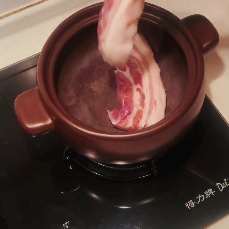 Rohe Schweinebauch-Streifen werden mit einer Metallzange in einen vorgeheizten dunklen Tontopf gelegt.