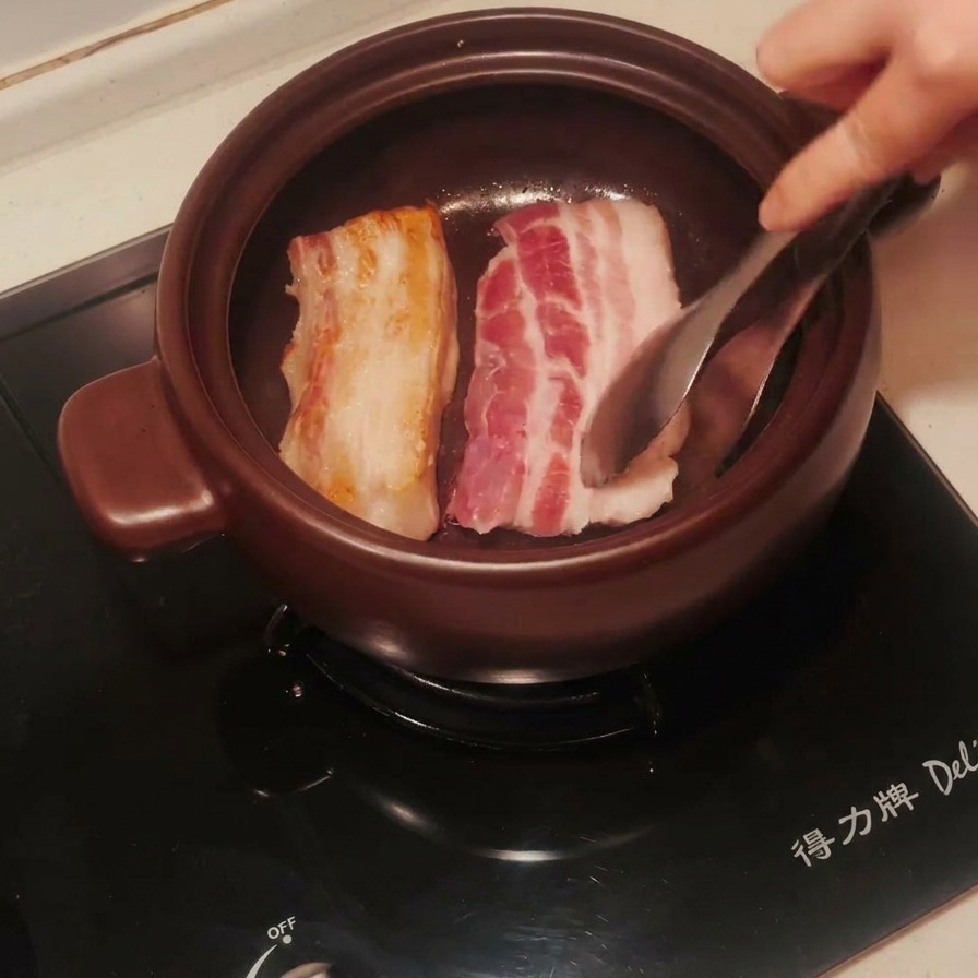 Schweinebauch-Streifen in einem Tontopf werden gewendet; eine Seite ist bereits perfekt goldbraun gebraten.