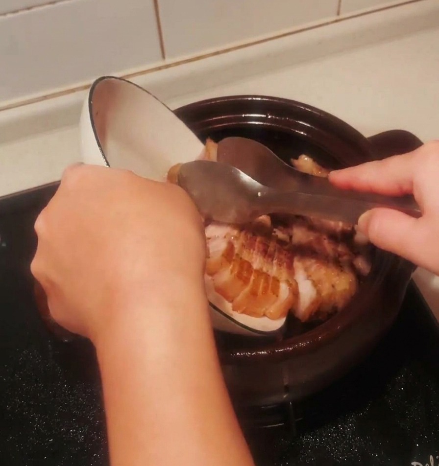 Angebratene Schweinebauchstücke werden aus einer weißen Schüssel zurück in den dunklen Tontopf geschüttet.