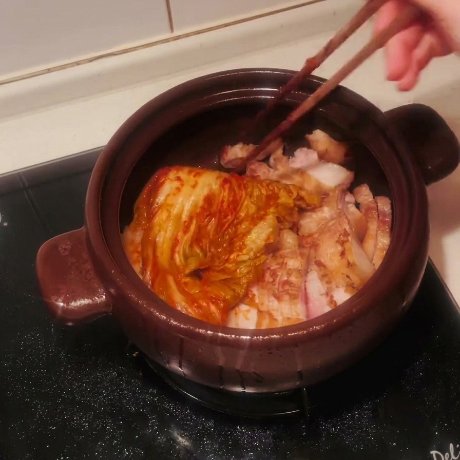 Ein ganzer Kopf gewürzter roter Kimchi wird mit Stäbchen über den Schweinebauch in den Tontopf gegeben.