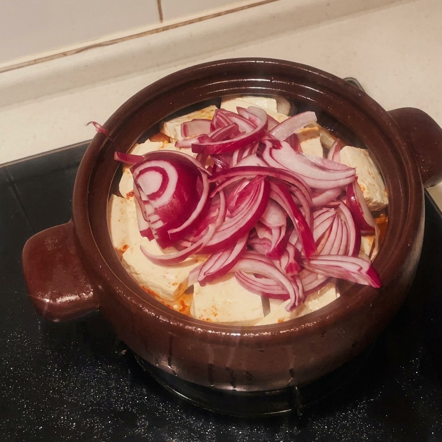 Eine Schicht aus geschnittenen roten Zwiebeln bedeckt den Tofu im Tontopf.