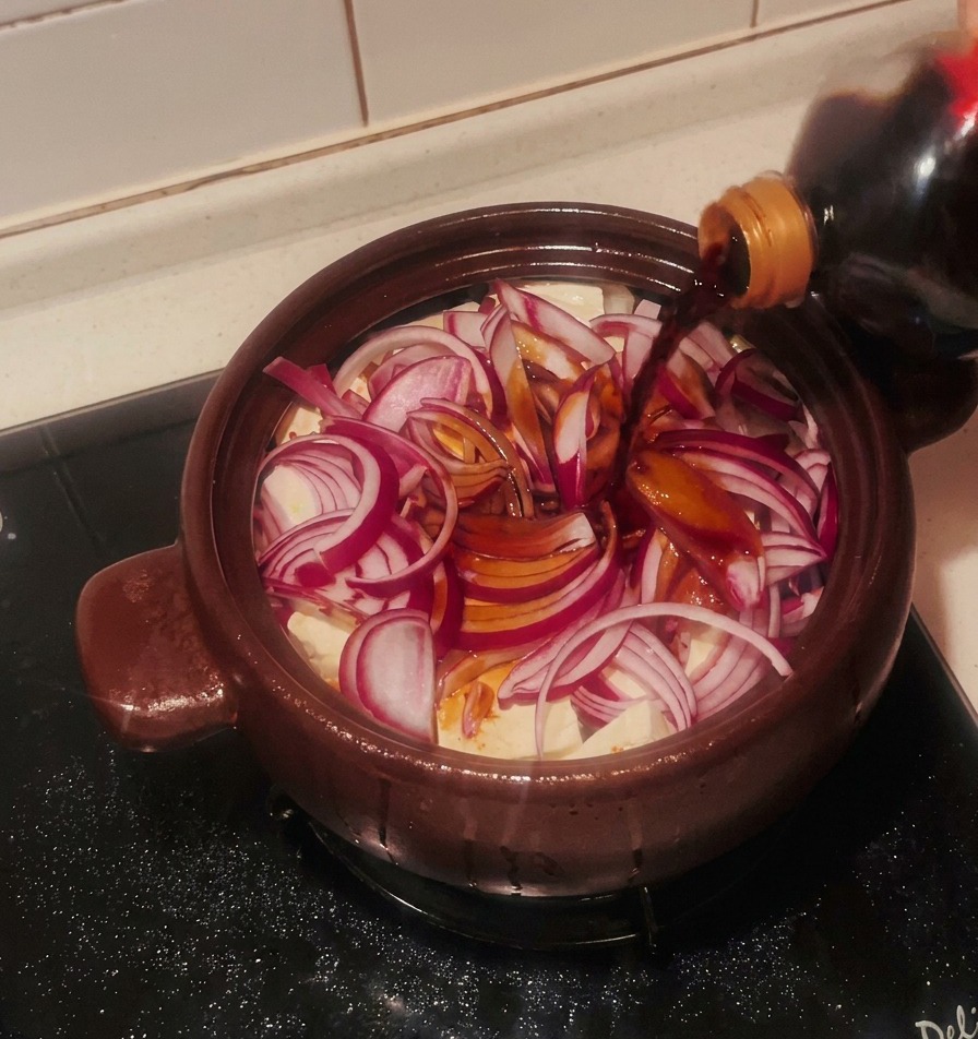 Dunkle Sojasauce wird über die roten Zwiebeln und den Tofu im braunen Tontopf gegossen.