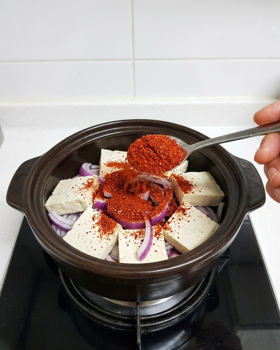 Ein Löffel gibt feines, leuchtend rotes Chilipulver auf den Tofu und die Zwiebeln im Topf.