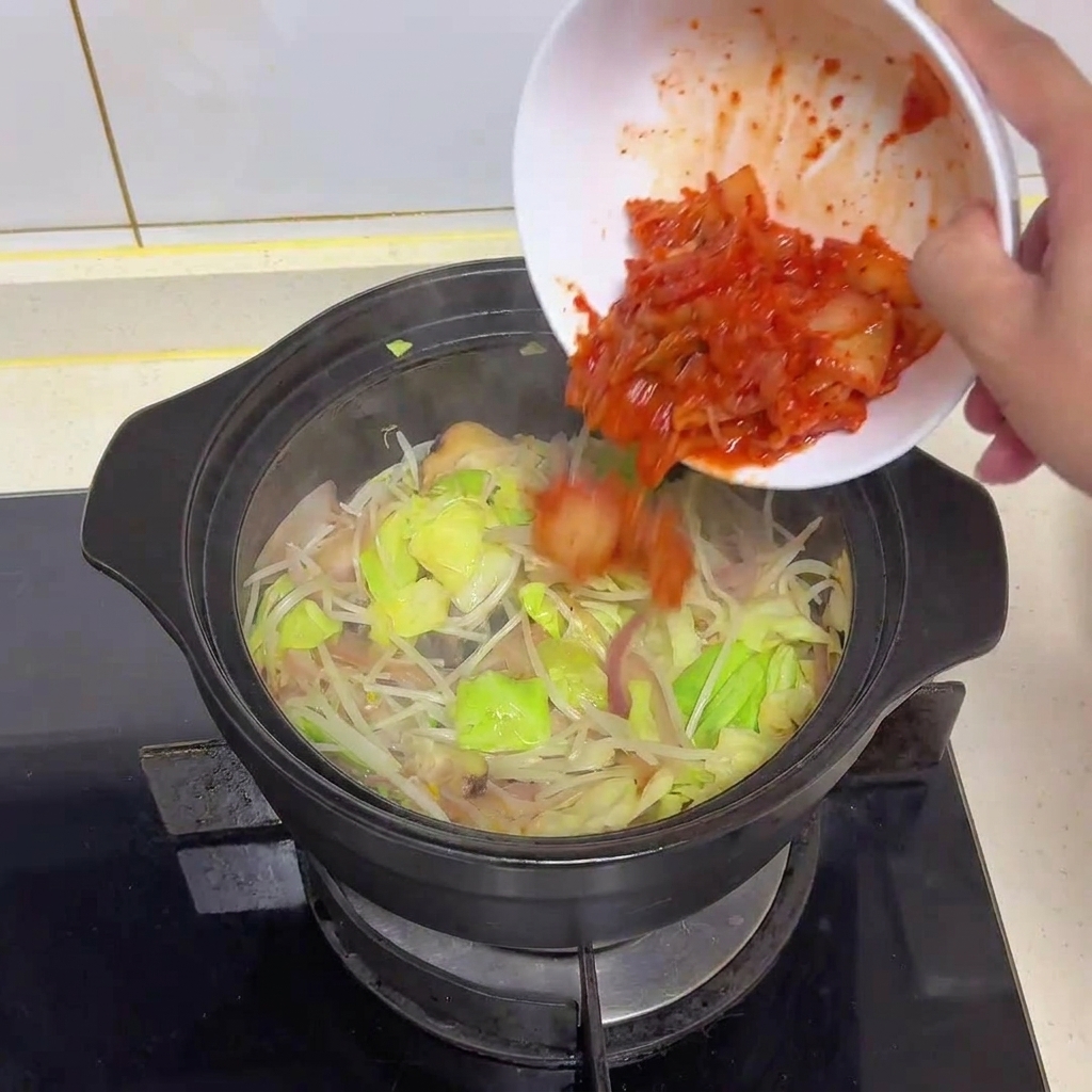 Leuchtend rotes, gehacktes Kimchi wird aus einer Schüssel in den Topf mit dem angebratenen Gemüse gegossen.