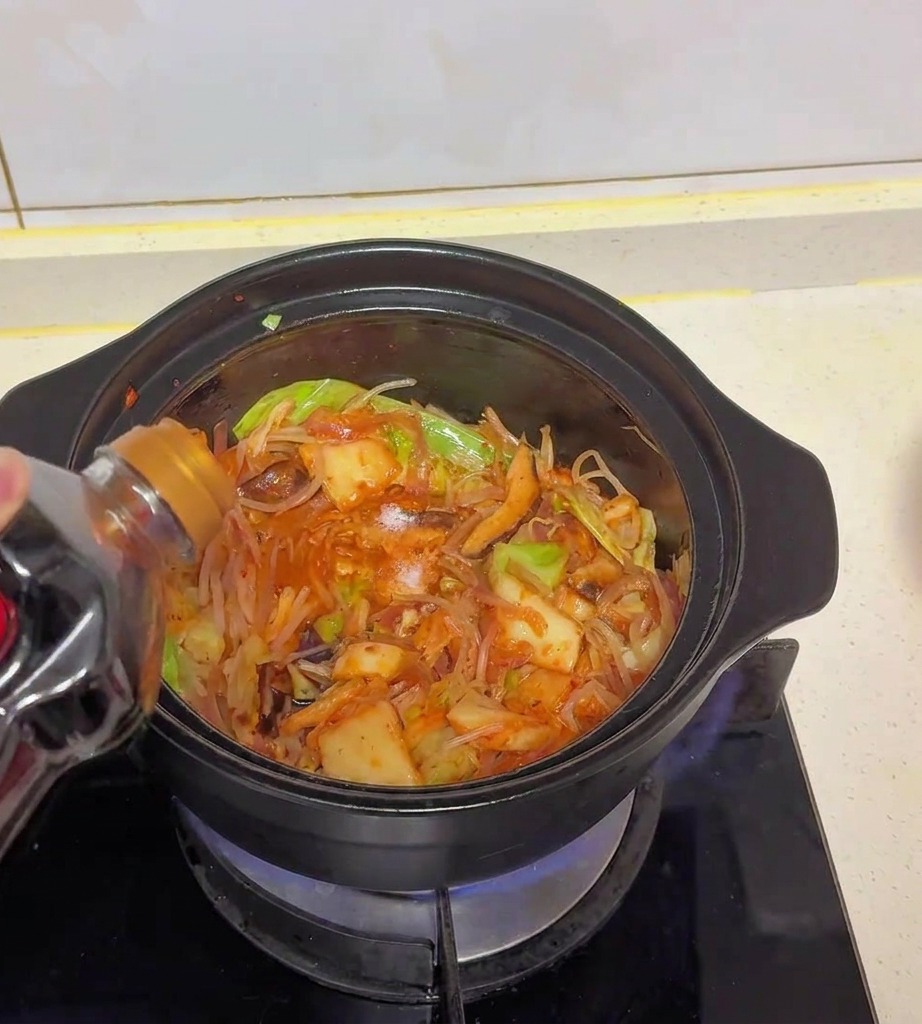 Helle Sojasauce wird aus einer dunklen Plastikflasche in einen Topf mit angebratenem Kohl, Kimchi und Pilzen gegossen.