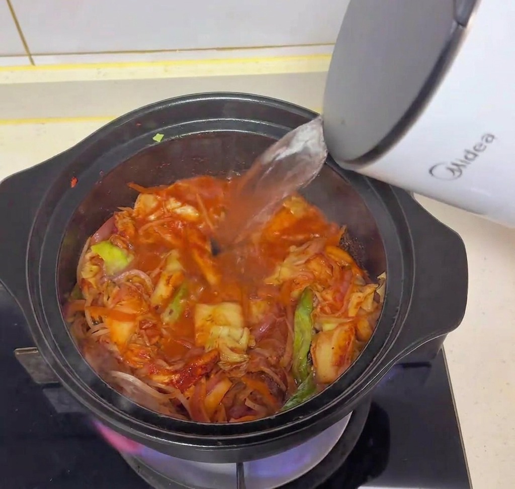 Klares, heißes Wasser wird aus einem weißen Wasserkocher in einen schwarzen Topf mit angebratenem Kimchi und Gemüse gegossen.