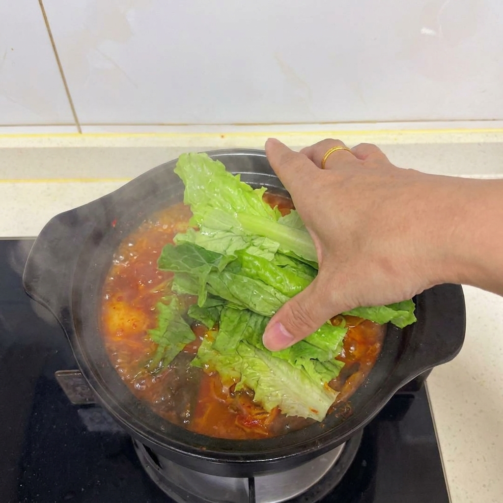 Eine Hand drapiert leuchtend grüne Salatblätter über einen dampfenden Topf mit roter Kimchi-Tofu-Suppe.