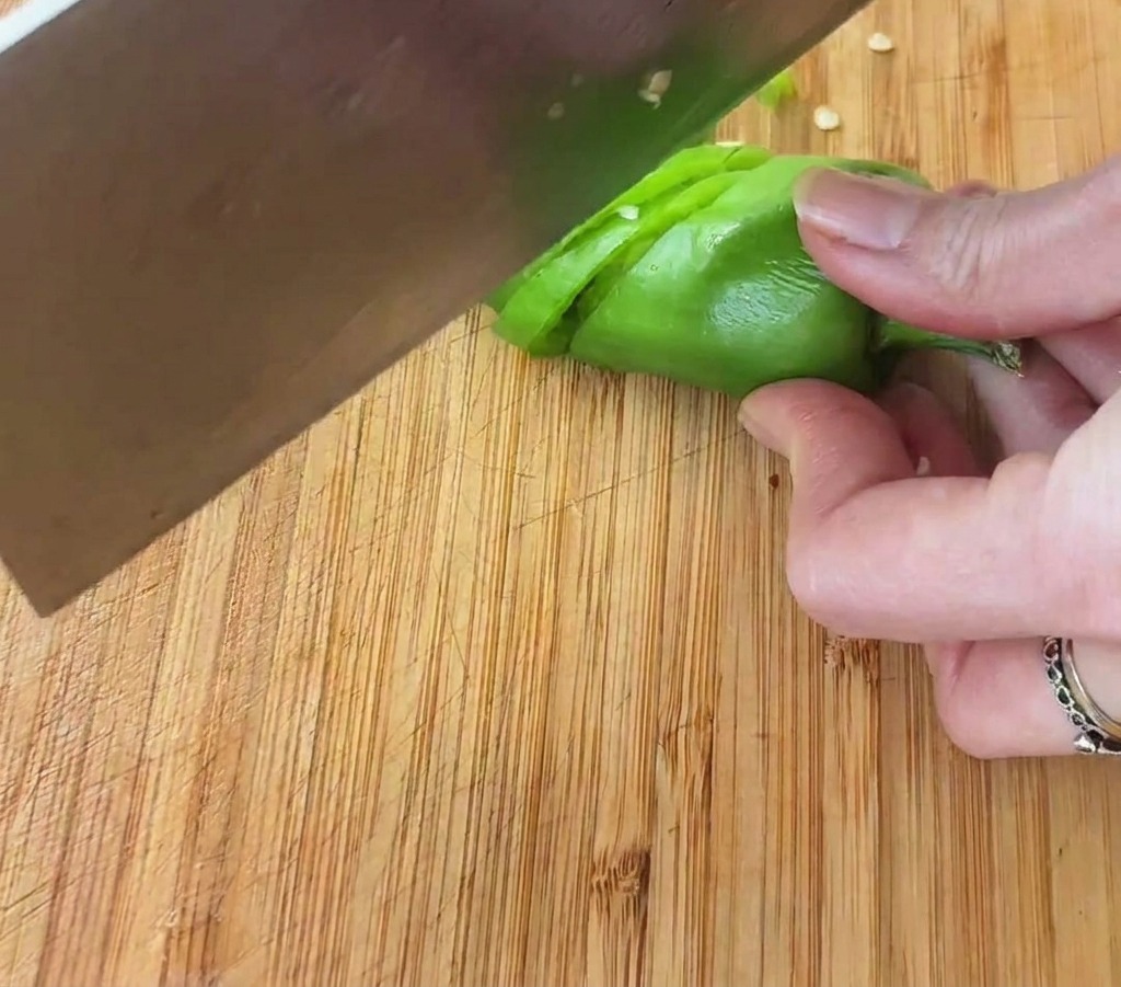 Eine Hand, die eine hellgrüne Chilischote mit einem großen Messer in kleine Ringe auf einem Holzbrett schneidet.