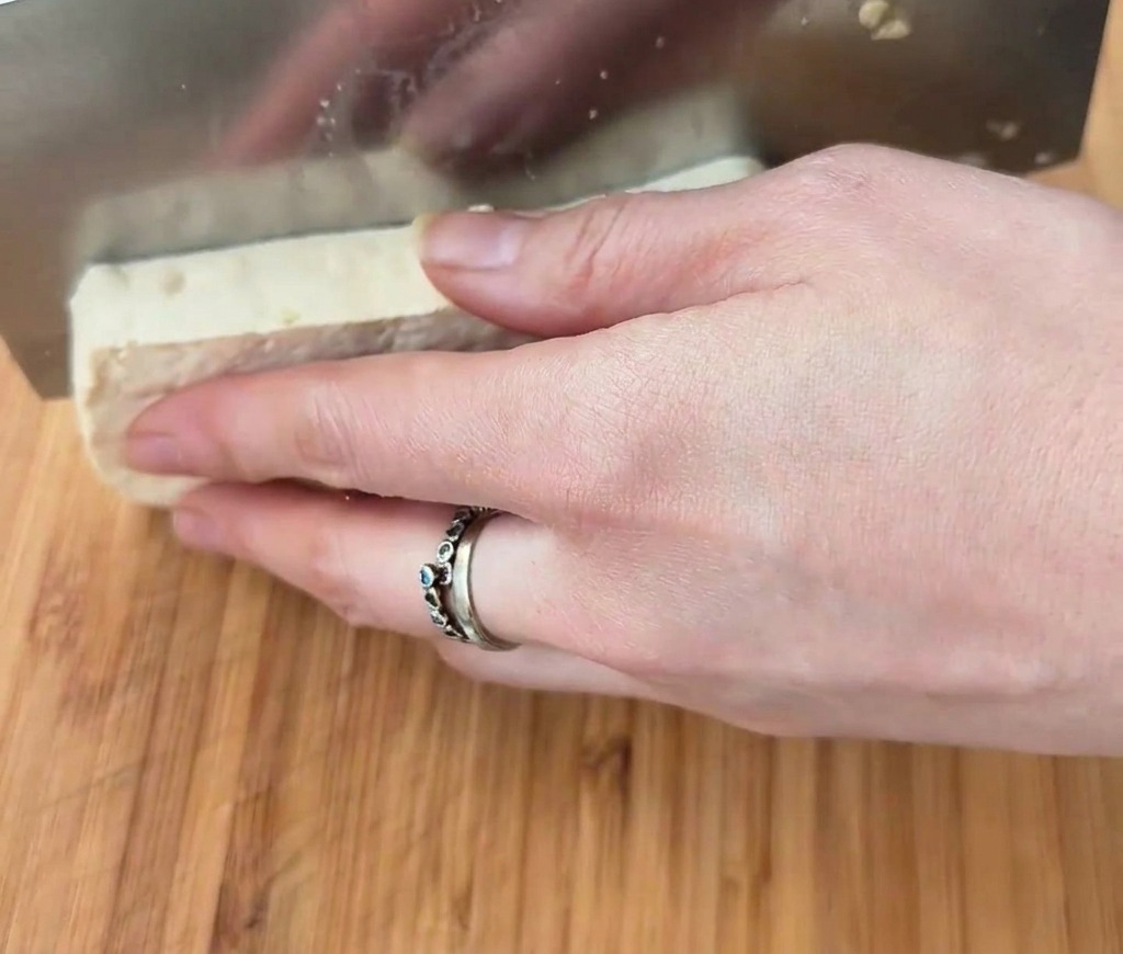 Ein rechteckiger Block weißer Tofu, der mit einem breiten Messer auf einer Holzoberfläche in Würfel geschnitten wird.
