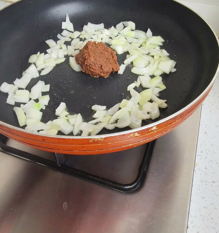 Ein Löffel dunkle Sojabohnenpaste in einer Pfanne mit bereits angedünsteten weißen Zwiebeln.