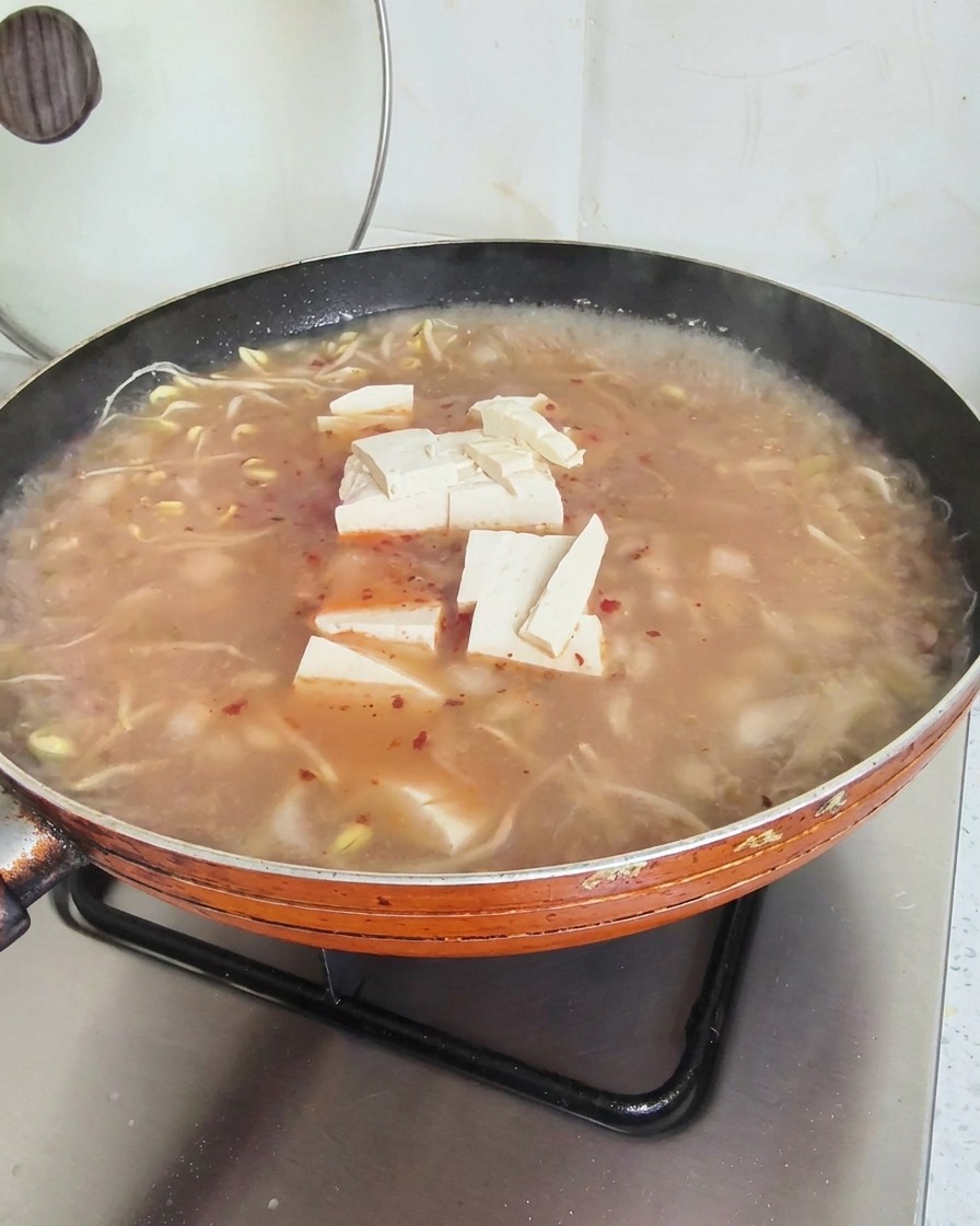 Tofuwürfel schwimmen auf der Oberfläche einer köchelnden, rötlichen Suppe in einer großen Pfanne.