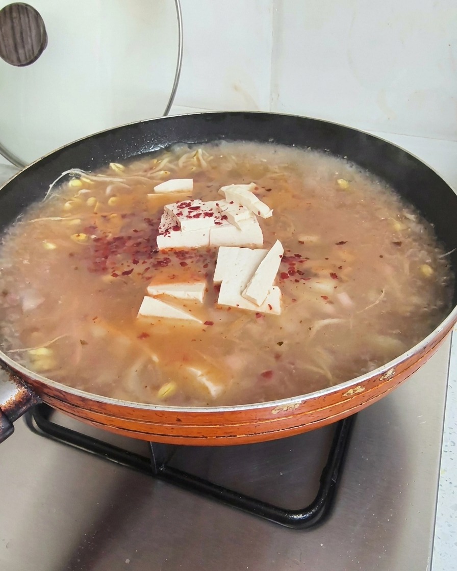 Leuchtend rote Chiliflocken sind über einer köchelnden Suppe mit Tofu und Sojasprossen verteilt.