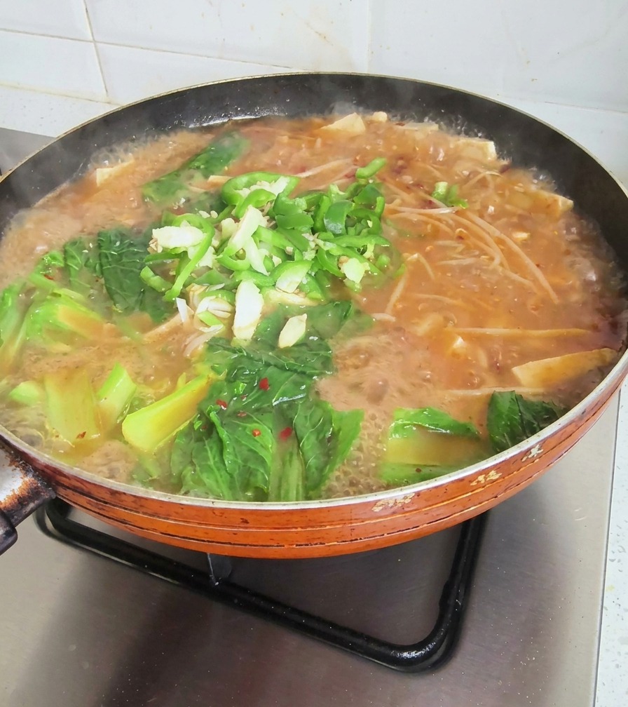 Eine köchelnde Pfanne mit koreanischer Suppe, garniert mit grünem Blattgemüse, Chiliringen und gehacktem Knoblauch.