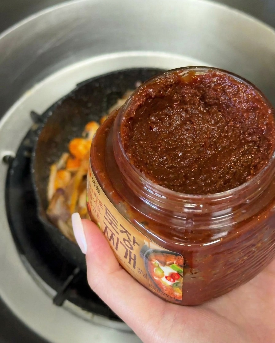 Ein großer Löffel dunkler koreanischer Sojabohnenpaste wird zum Gemüse in die Pfanne gegeben.