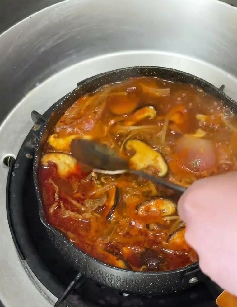 Ein Metalllöffel gießt einen Strahl bernsteinfarbenes Sesamöl in die köchelnde, scharfe Suppe.