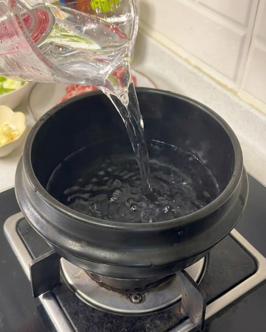 Wasser wird aus einem Messbecher in einen schwarzen Keramiktopf auf einem Gasherd gegossen