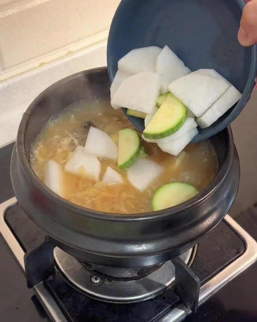 Rettich- und Zucchini-Scheiben werden aus einer Schale in die kochende braune Brühe gegeben
