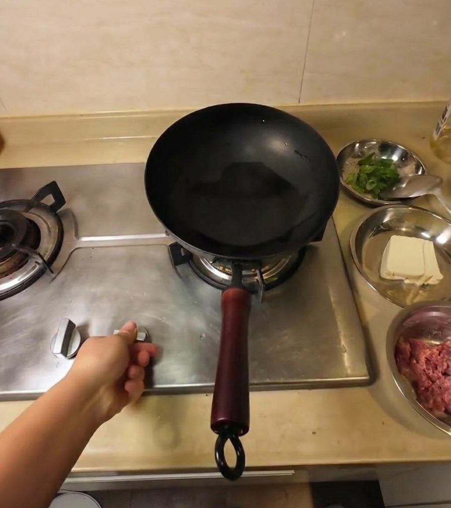 Eine Hand am Gasherd, die einen schwarzen Wok mit klarem Speiseöl erhitzt.
