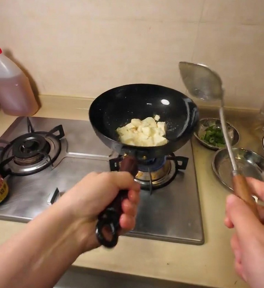Ein Metallspatel zerteilt den Tofu-Block im heißen Öl direkt im Wok in mundgerechte Stücke.