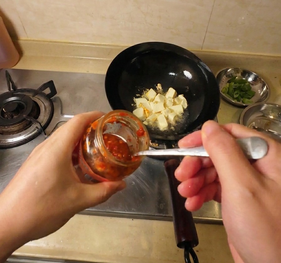 Ein Löffel leuchtend rote Chilisauce wird aus einem Glas zu den goldbraunen Tofustücken im Wok gegeben.