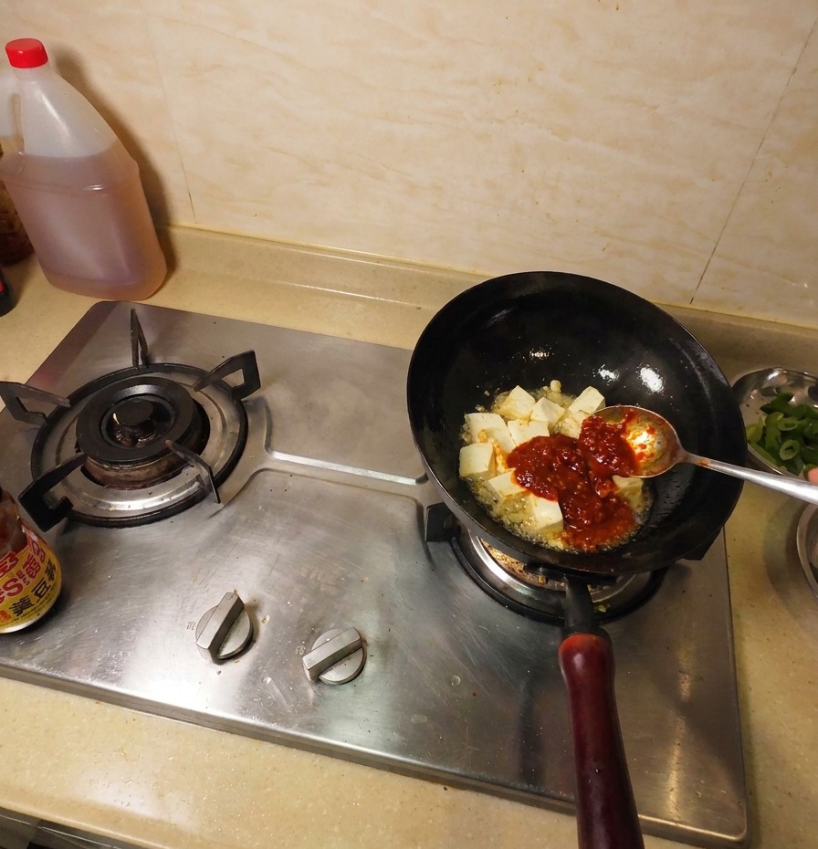 Dunkelbraune Sojabohnenpaste auf einem Metalllöffel wird zum Tofu und der Chilisauce in den Wok gegeben.