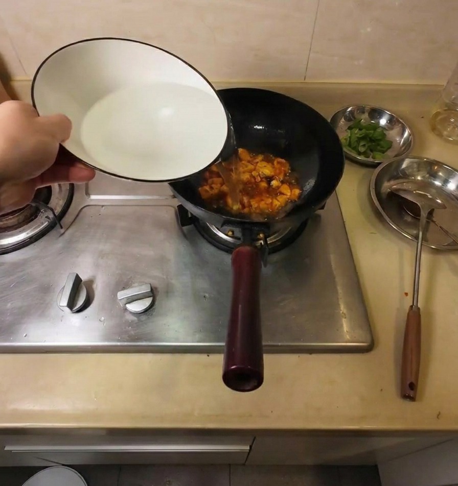 Heißes Wasser wird aus einer weißen Keramikschale in den Wok zu den Tofustücken und Gewürzen gegossen.