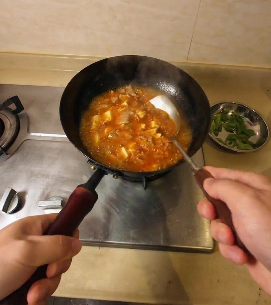 Rühren in einer köchelnden, sämigen orangeroten Suppe mit Tofu und Rindfleisch im schwarzen Wok.