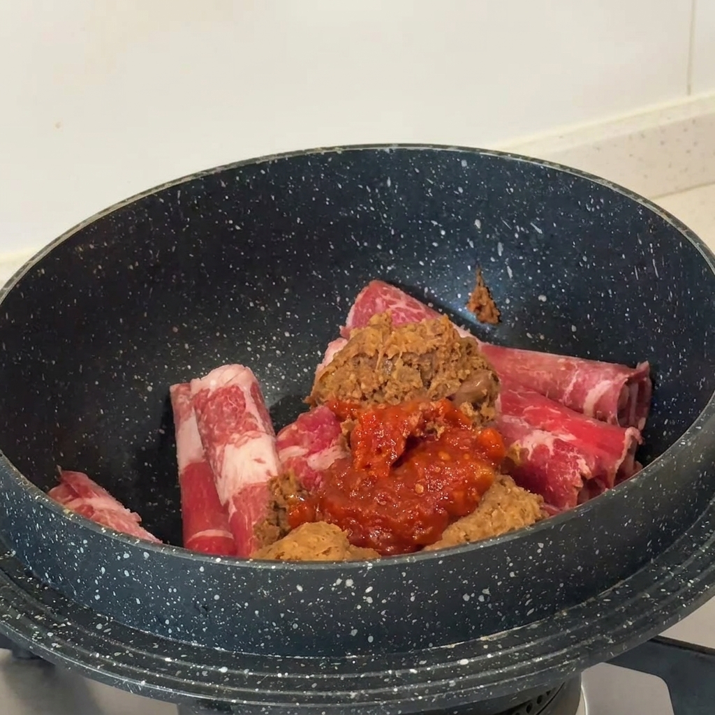 Häufchen aus brauner Sojabohnenpaste und leuchtend roter Chilipaste liegen auf rohen Fleischscheiben in einem dunklen Topf.