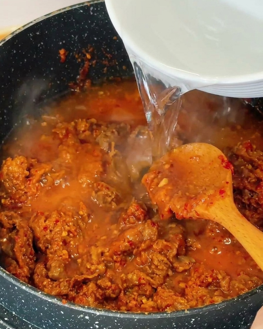 Klares Wasser wird aus einer weißen Schale in den Topf mit dem angebratenen Fleisch und der blubbernden Brühe gegossen.