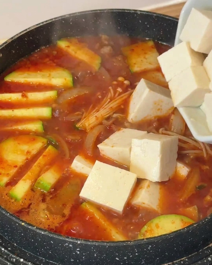 Weiße Tofuwürfel werden in einen Topf mit köchelnder Suppe, Zucchini und Enoki-Pilzen gegeben.