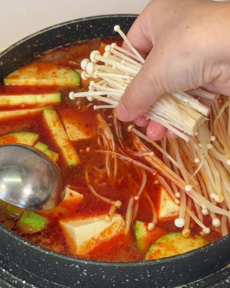Eine Hand legt ein Bündel langer, dünner Enoki-Pilze in die kochende Suppe mit Tofu und Zucchini.