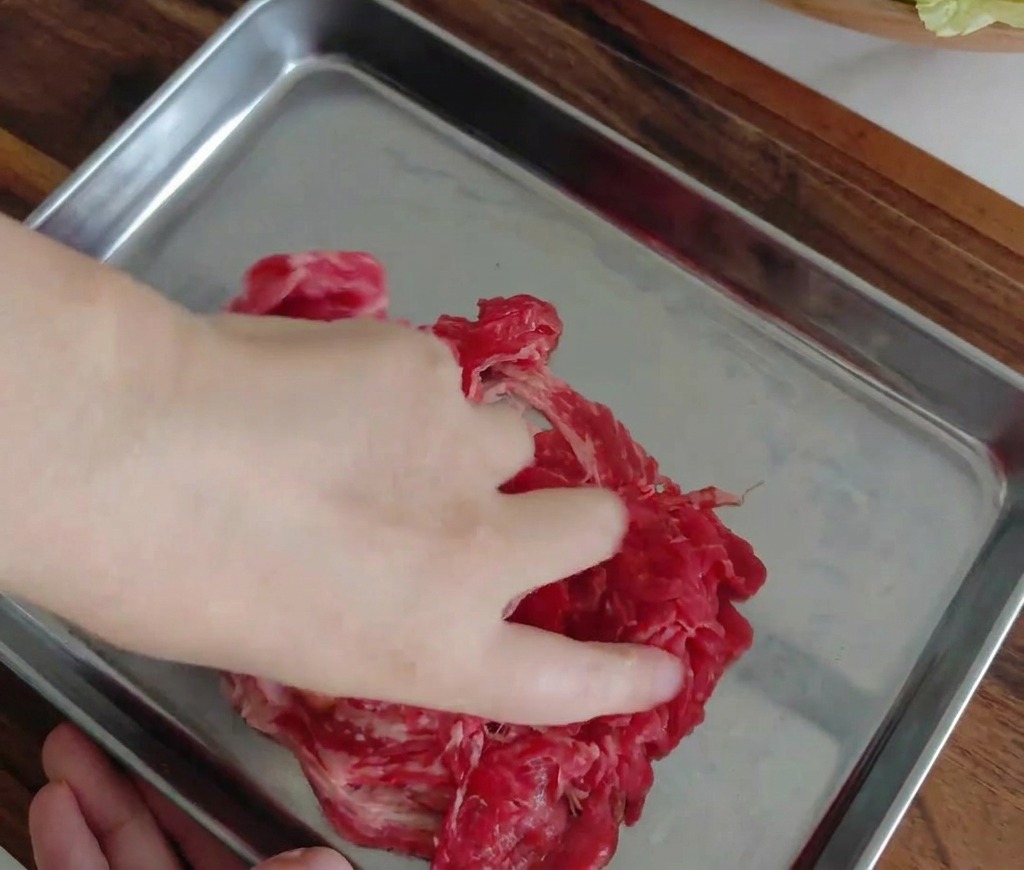 Eine Hand vermengt die marinierten Rindfleischscheiben auf einem Metalltablett.