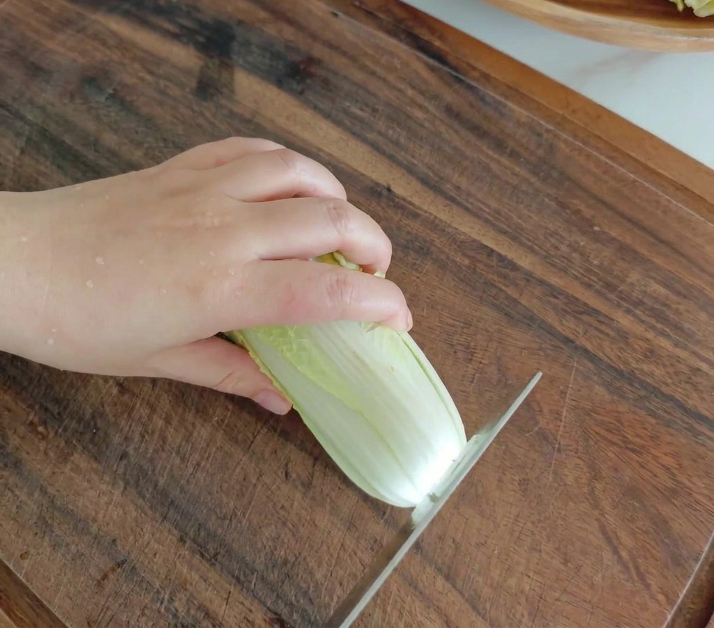 Ein Küchenmesser schneidet einen frischen Baby-Kohlkopf auf einem dunklen Holzschneidebrett in Stücke.
